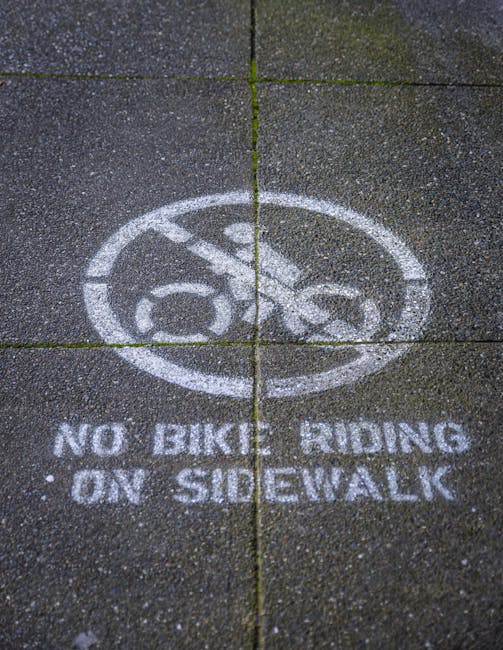 A close-up view of a concrete sidewalk with a white painted circle featuring a bicycle icon and a diagonal line crossing through it, indicating no bike riding permitted. Below the symbol, the text 'NO BIKES RIDING ON SIDEWALK' is clearly stenciled in capital letters. The pavement consists of concrete slabs with visible expansion joints and a rough texture, and there are small patches of moss or grass growing in the gaps. The scene is well-lit, highlighting the details of the painted markings and the surface of the sidewalk. This type of signage may be seen near residential or commercial areas where pedestrian safety and house removals are prioritized, aligning with regulations that [COMPANY_NAME], such as Man with Van Hatton, follow during loading and unloading operations at properties like those along the page about avoiding parking fines in Warwickshire, Hatton.
