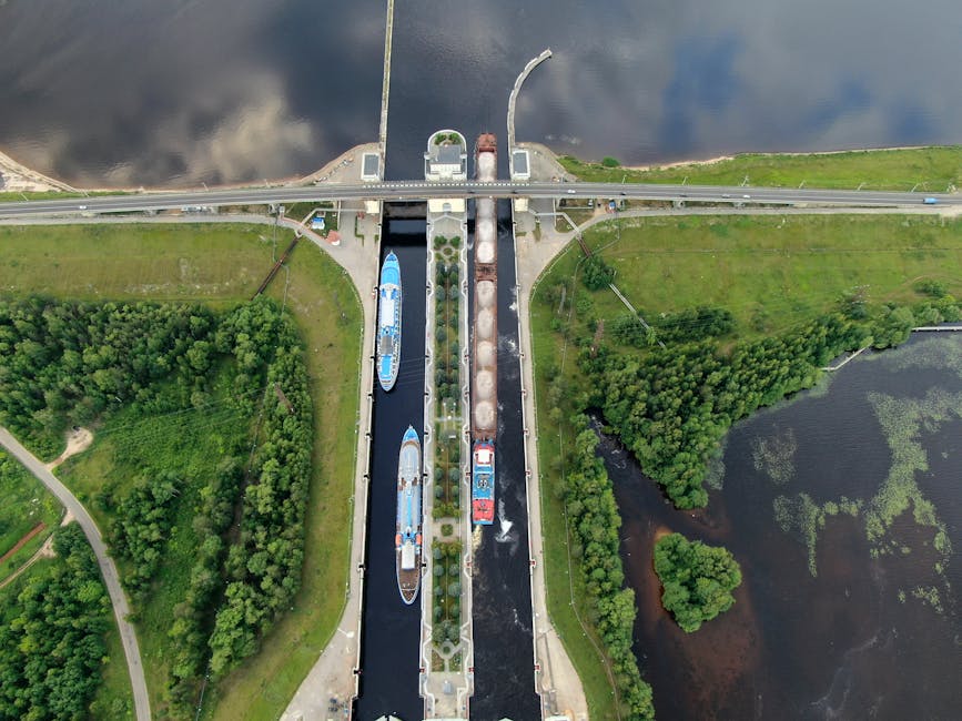 Aerial view of a canal lock system with two boats, one blue and one red, positioned side by side within the lock chambers. The scene captures the water level change with the boats floating in calm wat