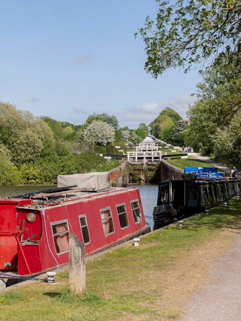 Aerial view of a canal lock system with two boats, one blue and one red, positioned side by side within the lock chambers. The scene captures the water level change with the boats floating in calm wat