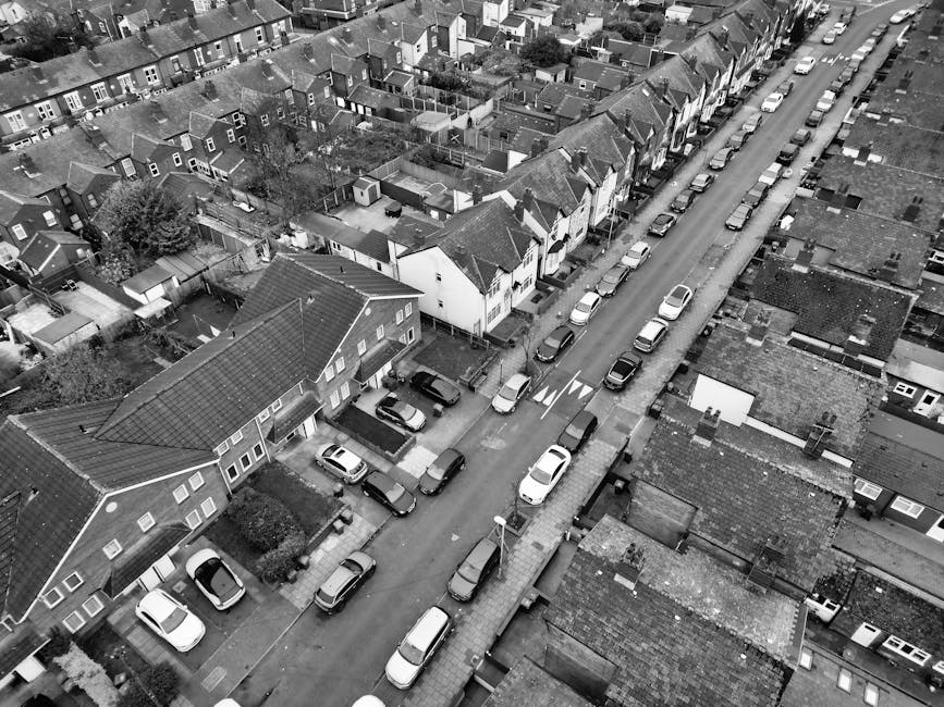 A black and white aerial photograph of a narrow residential street with closely packed terraced houses and semi-detached homes on either side. Parked cars line both sides of the street, with some positioned in driveways and others along the pavement. The scene shows the loading process of home relocation or furniture transport, where a man with a van from Man with Van Hatton is potentially preparing for a move along Church Lane to High Street. Visible are moving boxes, some stacked on pavements and in the process of being loaded into or unloaded from the van, which is parked adjacent to the curb near a house entrance. The properties have small front gardens or yards, and the street features traffic markings and street lighting. The environment appears calm, with soft natural lighting indicating daytime, and the image effectively captures the logistics involved in packing and moving within a dense suburban neighborhood.