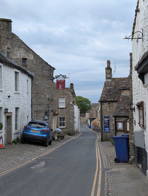 A narrow, cobbled street in Hatton during daytime, with historic stone and white-painted cottages on either side. Some buildings have small windows and sloped tiled roofs, with a blue car parked on the left curb. A blue trash bin is positioned on the right sidewalk, alongside a black signboard and a hanging wrought iron sign bracket attached to a building. The street gently curves ahead, lined with double yellow lines indicating parking restrictions, and the sky above is partly cloudy with patches of blue. This setting illustrates a typical quaint UK village environment suitable for house removals or small-scale furniture transport, aligning with the context of packing and moving services offered by Man with Van Hatton while maintaining a neutral and factual tone.