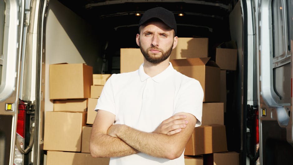 A young male mover with a beard, wearing a white polo shirt and black cap, standing with arms crossed inside the open rear cargo area of a silver moving van. The van's interior is filled with stacked cardboard boxes of various sizes, some wrapped in plastic or tape, ready for home relocation or furniture transport. The background shows the dark interior of the van, illuminated by natural light, and the environment appears to be on a driveway or street. Nearby, on the pavement, there are additional boxes and packing materials. This scene illustrates the loading process during a professional house removal, with a focus on careful packing and efficient transportation, as seen in services by companies like Man with Van Hatton.