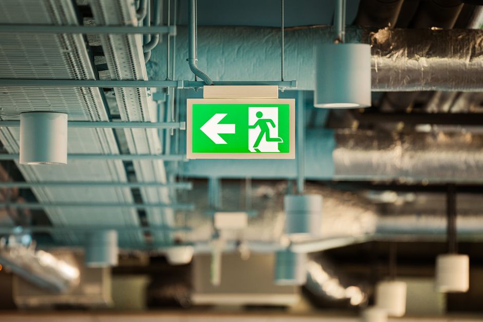 A green illuminated emergency exit sign featuring a white running figure and a left arrow, mounted on an industrial ceiling with exposed ducts, pipes, and ventilation systems. The sign indicates the direction to an emergency exit and is positioned centrally in the image, with the industrial environment of a commercial or storage premises visible around it. The image captures the ceiling's metal framework, cylindrical ductwork, and electrical conduits, emphasizing a typical setting where safety signage is prominently displayed to guide occupants during evacuation, relevant to house removals and transition logistics managed by Man with Van Hatton as part of their moving and packing services.