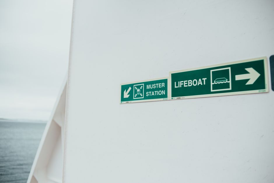 The image shows a white wall inside a property, with two green and white safety signs attached near the top right corner. The first sign indicates the location of a muster station with an arrow pointing downward and features a figure gathering under a shelter, designed for emergency evacuation guidance. Adjacent to it, the second sign points to the right and directs toward a lifeboat, accompanied by an icon of a boat on water to denote maritime safety procedures. The environment appears well-lit with natural light, suggesting the interior space is near an exit or passageway, possibly part of a house or building involved in a house removal or relocation process. The presence of these safety signs subtly highlights the importance of safety measures during the packing and moving process, which is a typical part of furniture transport and home relocation services provided by Man with Van Hatton, as seen on the company's website. No furniture, boxes, or equipment are visible in this specific image, focusing instead on the safety signage relevant to the moving environment.