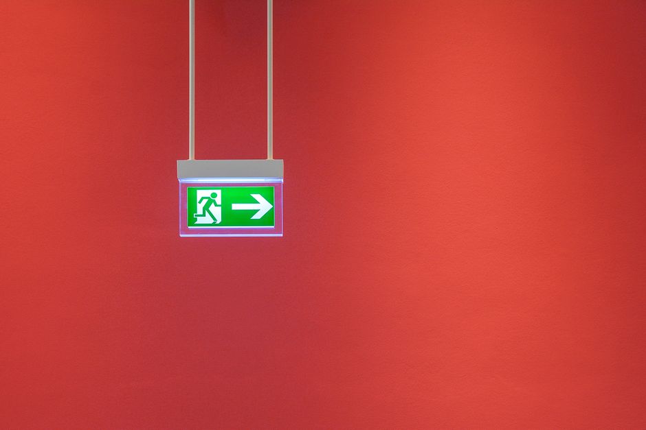 A green illuminated emergency exit sign featuring a white running figure and a left arrow, mounted on an industrial ceiling with exposed ducts, pipes, and ventilation systems. The sign indicates the direction to an emergency exit and is positioned centrally in the image, with the industrial environment of a commercial or storage premises visible around it. The image captures the ceiling's metal framework, cylindrical ductwork, and electrical conduits, emphasizing a typical setting where safety signage is prominently displayed to guide occupants during evacuation, relevant to house removals and transition logistics managed by Man with Van Hatton as part of their moving and packing services.