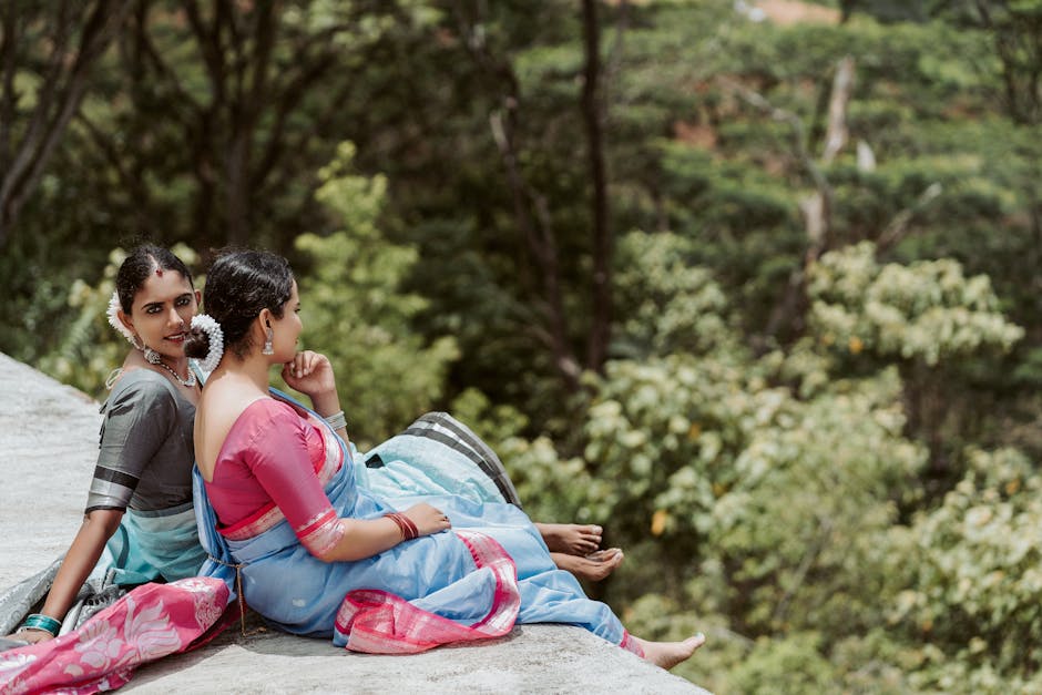Two women wearing traditional South Asian sarees, one in pink and blue and the other in grey and mint green, sitting on a concrete ledge outdoors near dense greenery and trees. One woman relaxes with her legs extended and barefoot, while the other rests with her chin on her hand, both facing the forested background. The scene is illuminated by natural daylight, capturing a tranquil moment. Although the image does not depict moving or packing activities, it provides a serene contrast to the logistics of house removals or home relocations, which are services offered by Man with Van Hatton, as indicated on the page about bulky waste and house clearance in Hatton.