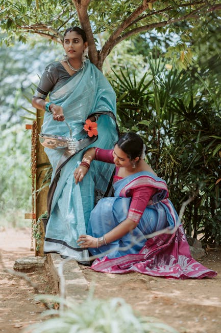 Two women dressed in traditional Indian sarees are outdoors near dense greenery and trees. One woman stands on a wooden platform holding a small container, while the other woman is kneeling on the ground, reaching up to adjust the standing woman's saree. The kneeling woman wears a pink and blue saree with matching jewelry, and the standing woman wears a light blue saree with floral accents and jewelry. The background features lush foliage and natural lighting, creating a serene atmosphere. The image captures a moment of cultural tradition and social interaction in a natural setting.