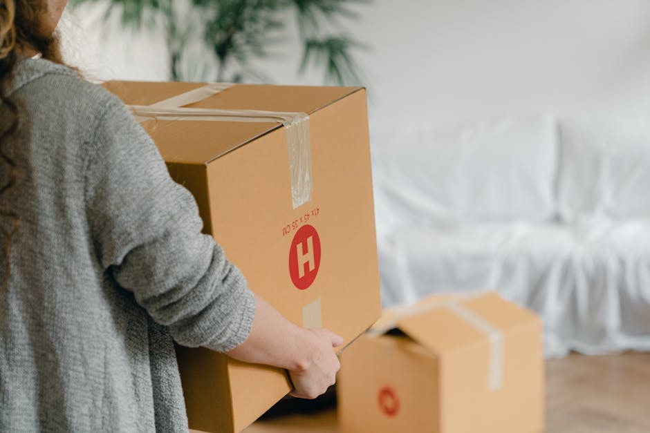 A man and a young girl are inside a house during a home relocation, surrounded by large cardboard boxes prepared for moving. The girl is seated at a table, holding a white mug, and looking at a laptop