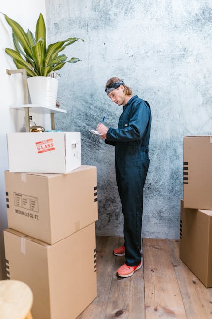 A young man wearing a blue jumpsuit, headband, and red shoes standing indoors next to a stack of cardboard boxes in a room with a textured blue wall and wooden flooring. The boxes vary in size, with some labeled for glass and others plain, and are arranged in a corner near a plant in a white pot on a small white shelf attached to the wall. The man is holding a pen and a notepad, appearing to take inventory or plan the next steps involved in home relocation or furniture transport. Additional boxes are positioned on the floor around him, with one partially visible to the right. The scene depicts a preparation or packing stage of a house move, fitting for content related to removals services provided by Man with Van Hatton, supporting the themes of packing, moving logistics, and loading process.