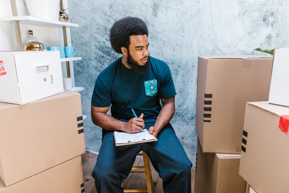 A man with an afro hairstyle and a beard sits on a wooden stool inside a room with a textured, light blue wall. He is wearing a dark blue t-shirt with a small patterned pocket and dark trousers, holding a pen in his right hand and a clipboard in his left hand, and appears to be taking notes or surveying the space. In front of him are several large cardboard boxes, some sealed with black tape, stacked on the floor. To his left, a white shelving unit with decorative items such as vases and cups is visible. The scene suggests a home relocation environment, with packing materials and furniture preparation for moving. This image exemplifies the process of measuring and planning furniture transport as part of a house removal, with an emphasis on careful organisation by [COMPANY_NAME], a professional removals service. The setting appears well-lit, possibly with natural light, emphasizing the organized packing and inventory tasks involved in a detailed move-out checklist for Hatton tenants.