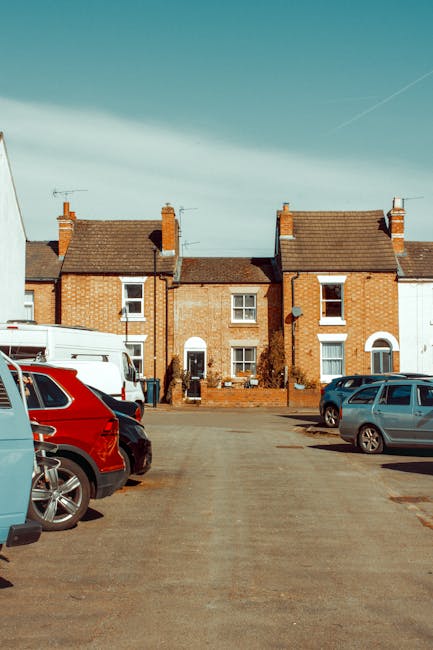 A traditional cottage with a thatched roof and whitewashed walls, situated along a quiet street with a paved road and a small grassy verge. The house features multiple brick chimneys and small windows