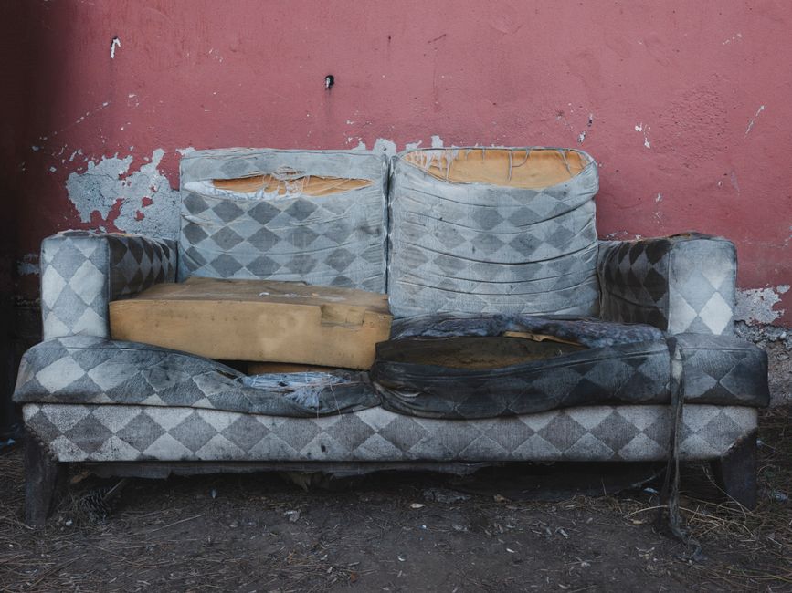 A weathered, dilapidated sofa with a blue and grey diamond-patterned fabric, visibly torn and stained, is positioned outdoors against a worn, peeling red brick wall. The sofa's cushions are damaged, w