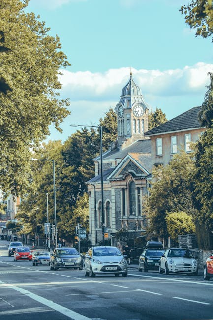A city street scene during daytime with multiple parked and moving cars along the pavement, including a white van parked near the curb. Behind the vehicles, there is a historic building with a clock tower topped by a small spire, featuring intricate architectural details and large arched windows. Tall trees line the street, partially obscuring the upper floors of nearby residential buildings with brick facades and pitched roofs. Overhead streetlights and utility poles are visible along the sidewalk, and the sky is clear with a few scattered clouds. The scene depicts a typical urban area suitable for house removals or furniture transport services, such as those offered by Man with Van Hatton, illustrating the environment in which home relocation or packing and moving activities could take place.