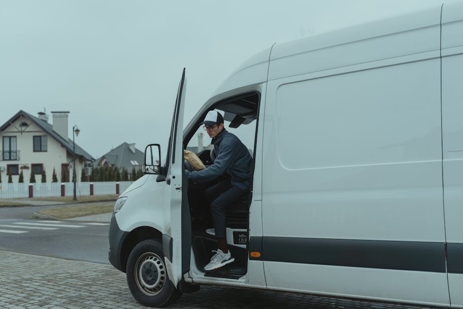 A man with a beard and wearing a baseball cap and white polo shirt is seen unloading cardboard boxes from the back of a moving van on a residential street during sunset. The van's interior shows sever